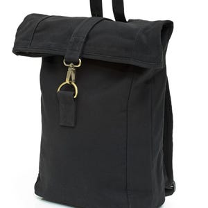 May include: A black canvas roll-top backpack featuring a top handle and a central vertical strap secured with a brass-toned metal lobster clasp and D-ring hardware. The bag has a rectangular structure with a folded top closure and adjustable straps.