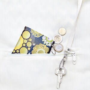 Op de afbeelding: Een klein, zwart-geel gepatroneerd muntportemonnee met een ritssluiting is gestopt in een witte ritssluiting. Een zilveren sleutelhanger met een sleutel is bevestigd aan de ritssluiting. Verschillende euro-munten liggen verspreid rond het portemonnee.