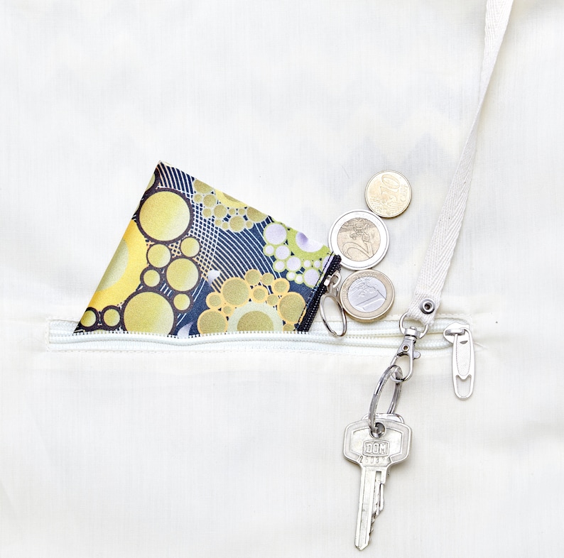 Op de afbeelding: Een klein, zwart-geel gepatroneerd muntportemonnee met een ritssluiting is gestopt in een witte ritssluiting. Een zilveren sleutelhanger met een sleutel is bevestigd aan de ritssluiting. Verschillende euro-munten liggen verspreid rond het portemonnee.