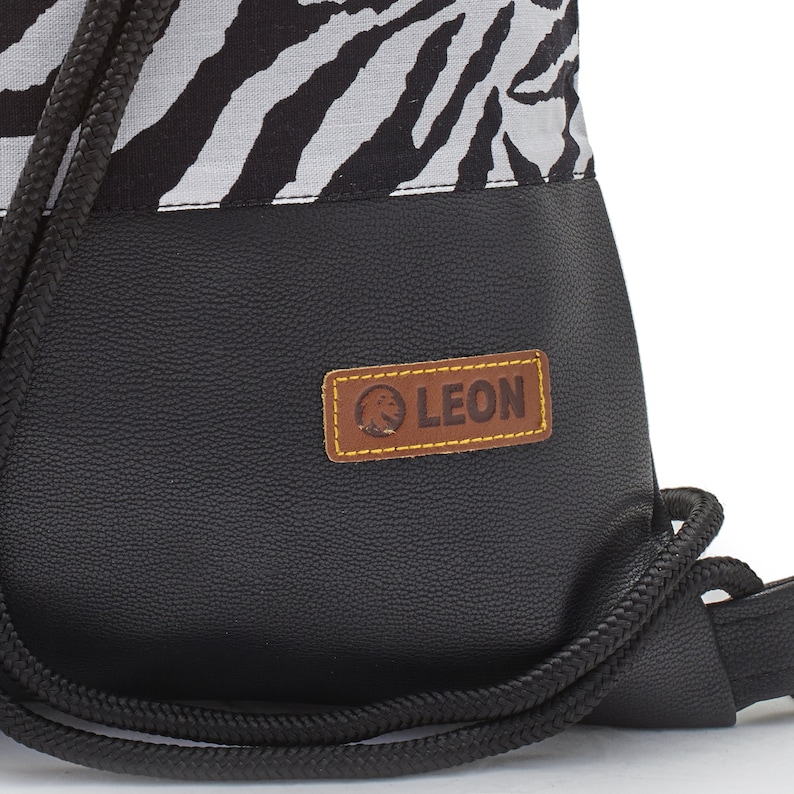 Op de afbeelding: Zwarte trekkoordtas met een zebraprint bovenkant en een zwarte lederen onderkant. De tas heeft een bruin lederen patch met het woord "LEON" in witte tekst.