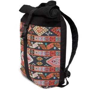 May include: A roll-top backpack featuring a black canvas upper section and a central panel with a multi-colored woven geometric pattern in shades of orange, teal, pink, and white. The bag includes a black vertical strap with a plastic buckle closure.