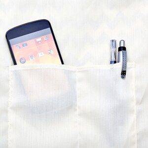 Op de afbeelding: Een wit overhemd met een zak waarin een zwarte smartphone en twee pennen zitten. De smartphone heeft een helder oranje en geel scherm met pictogrammen.