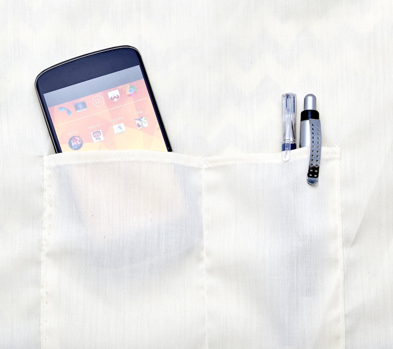 Op de afbeelding: Een wit overhemd met een zak waarin een zwarte smartphone en twee pennen zitten. De smartphone heeft een helder oranje en geel scherm met pictogrammen.