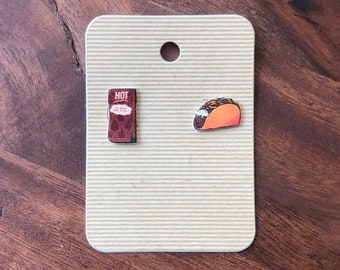 May include: A pair of food-themed earrings on a tan card. One earring is a small, brown packet with the word "HOT" and text. The other is a miniature taco with orange and brown details. The card has a hole at the top.
