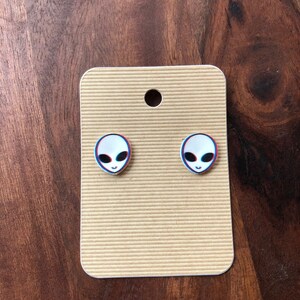 May include: A pair of silver stud earrings with a white alien face design. The alien face has blue and red 3D effects.