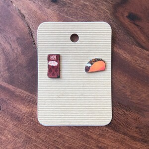 May include: A pair of food-themed earrings on a tan card. One earring is a small, brown packet with the word "HOT" and text. The other is a miniature taco with orange and brown details. The card has a hole at the top.