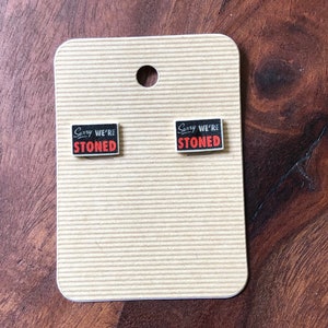 May include: A pair of black and red square earrings with the text "Sorry We're Stoned" on each earring.