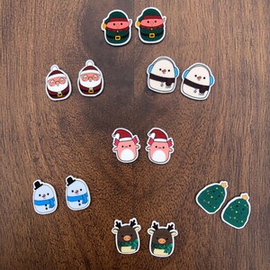 May include: A set of 12 Christmas-themed resin ornaments. The ornaments feature various characters, including Father Christmas, snowmen, elves, and axolotls. The ornaments are all in a cartoon style and are painted in bright colours.