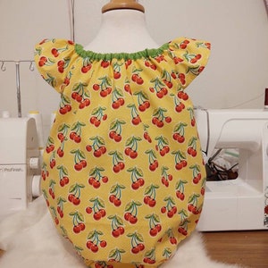 May include: A yellow baby onesie with a cherry print and green trim around the neckline. The onesie has short sleeves and a gathered neckline.
