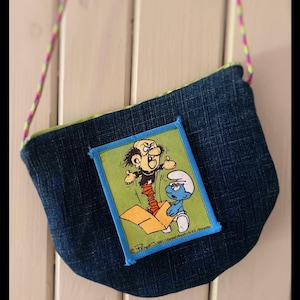 Könnte beinhalten: Eine Jeans-Geldbörse mit einem bunten Riemen. Die Geldbörse hat einen Aufnäher mit einem Cartoon-Bild eines mürrischen Charakters und eines Schlümpfes. Die Geldbörse hat ein grünes Futter und einen blauen Rand um den Aufnäher.