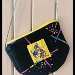 Könnte beinhalten: Schwarze Umhängetasche mit gelbem Reißverschluss und buntem Riemen. Die Tasche hat ein gelbes quadratisches Patch mit einem Cartoon-Bild von Tom und Jerry. Die Tasche hat einen schwarzen Hintergrund mit bunten Farbspritzern.