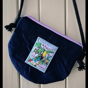 Könnte beinhalten: Dunkelblaue Denim-Umhängetasche mit hellviolettem Reißverschluss und Riemen. Die Tasche hat einen farbenfrohen Aufnäher mit einem Tukan, Schmetterlingen und dem Text "POLYNESIAN WILDLIFE". Die Tasche hat einen schwarzen Seilriemen.