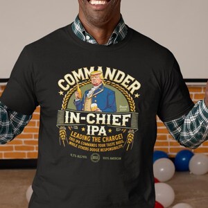 May include: A black t-shirt with the words "Commander In-Chief IPA" and an illustration of a man in a blue coat. The shirt is worn by a smiling person making a peace sign. American flags and red, white, and blue decorations are in the background.