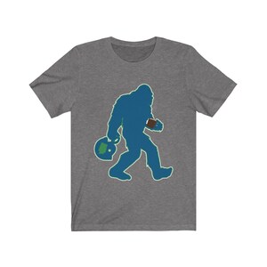 K&ouml;nnte beinhalten: Ein heidefarbenes T-Shirt mit einer blauen Silhouette von Bigfoot, der einen Football und einen gr&uuml;n-blauen Footballhelm tr&auml;gt.