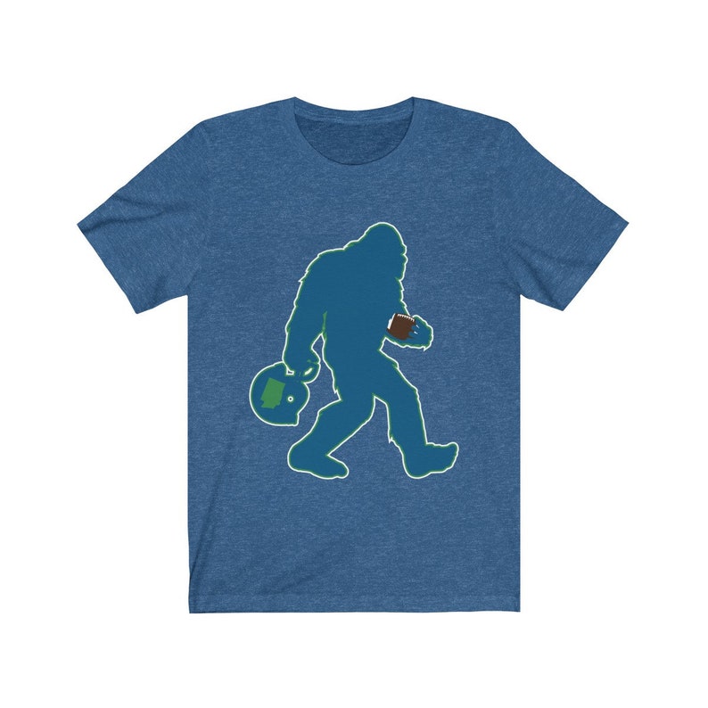 K&ouml;nnte beinhalten: Ein blaues T-Shirt mit einer gr&uuml;n-blauen Silhouette von Bigfoot, der einen Football und einen Helm tr&auml;gt.