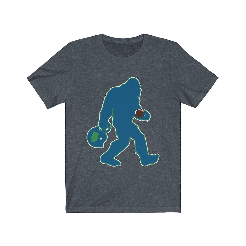 K&ouml;nnte beinhalten: Ein dunkelblaues T-Shirt mit einer gr&uuml;n-blauen Silhouette von Bigfoot, der einen Football und einen Helm tr&auml;gt.