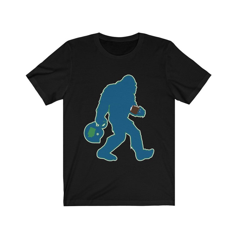 K&ouml;nnte beinhalten: Ein schwarzes T-Shirt mit einer blauen Silhouette von Bigfoot, der einen Football und einen Helm tr&auml;gt. Der Helm hat einen gr&uuml;nen Umriss und einen gr&uuml;nen Bundesstaat Washington im Inneren.