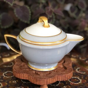 May include: A small gray ceramic creamer with a gold trim and a white lid. The creamer is sitting on a wooden coaster with a decorative design. The coaster is on a tray with a colorful pattern.