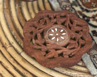 May include: A brown wooden coaster with an intricate carved design and a white inlay in the center. The coaster is sitting on a woven bamboo surface.