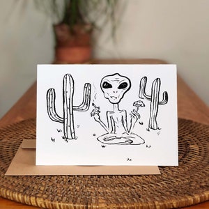May include: Black and white illustration of an alien sitting in a puddle with two cacti behind it. The alien is smoking a cigarette and holding a mushroom in its other hand.