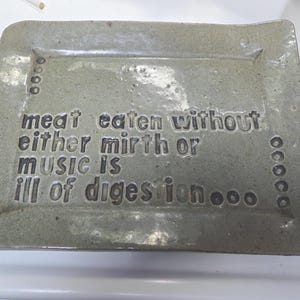 May include: A rectangular, speckled gray ceramic tray with a glossy finish. The surface features the embossed text: meat eaten without either mirth or music is ill of digestion, accompanied by small circular indentations along the left and right sides.
