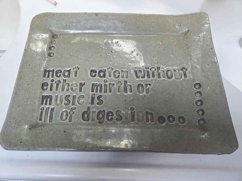 May include: A rectangular, speckled gray ceramic tray with a glossy finish. The surface features the embossed text: meat eaten without either mirth or music is ill of digestion, accompanied by small circular indentations along the left and right sides.