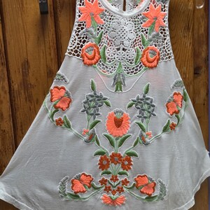 May include: White sleeveless top with intricate floral embroidery in coral, green, and gray. The top features a crochet neckline and a flowing, A-line silhouette, perfect for warm weather.