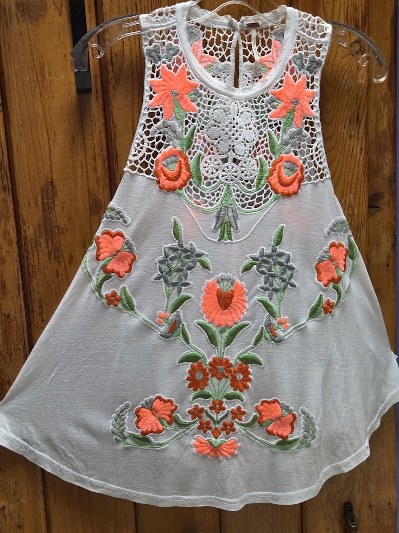 May include: White sleeveless top with intricate floral embroidery in coral, green, and gray. The top features a crochet neckline and a flowing, A-line silhouette, perfect for warm weather.