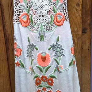 May include: White sleeveless top with intricate floral embroidery in coral, green, and gray. The top features a crochet neckline and floral patterns cascading down the front. The design is feminine and detailed, perfect for warm weather.