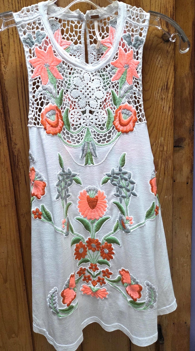 May include: White sleeveless top with intricate floral embroidery in coral, green, and gray. The top features a crochet neckline and floral patterns cascading down the front. The design is feminine and detailed, perfect for warm weather.