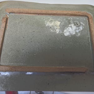 May include: A rectangular ceramic tray with a speckled sage green glaze and a raised, unglazed terracotta-colored rim on the underside. The surface features a glossy finish with visible dark flecks and a small, stamped maker's mark in one corner.