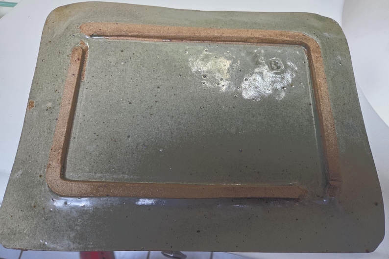 May include: A rectangular ceramic tray with a speckled sage green glaze and a raised, unglazed terracotta-colored rim on the underside. The surface features a glossy finish with visible dark flecks and a small, stamped maker's mark in one corner.