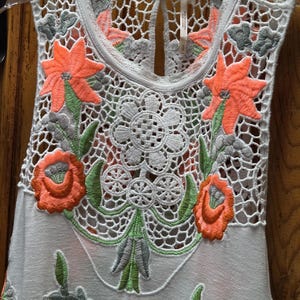 May include: A sleeveless white top with intricate floral embroidery. The top features a delicate crochet neckline and bodice, adorned with vibrant orange and green embroidered flowers and leaves. The design includes a keyhole back.