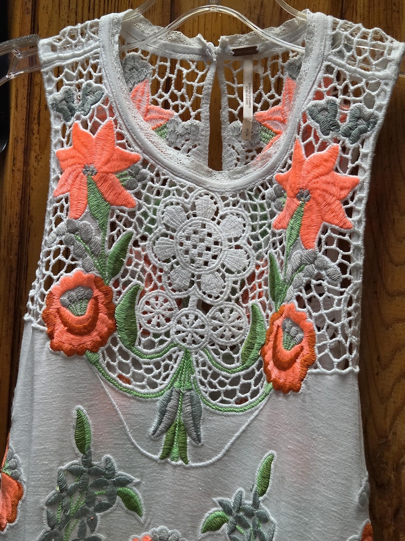 May include: A sleeveless white top with intricate floral embroidery. The top features a delicate crochet neckline and bodice, adorned with vibrant orange and green embroidered flowers and leaves. The design includes a keyhole back.