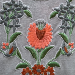 May include: Close-up of a white fabric with intricate floral embroidery. The design features orange, gray, and green flowers and leaves. The embroidery is detailed, with a textured appearance, creating a decorative pattern.