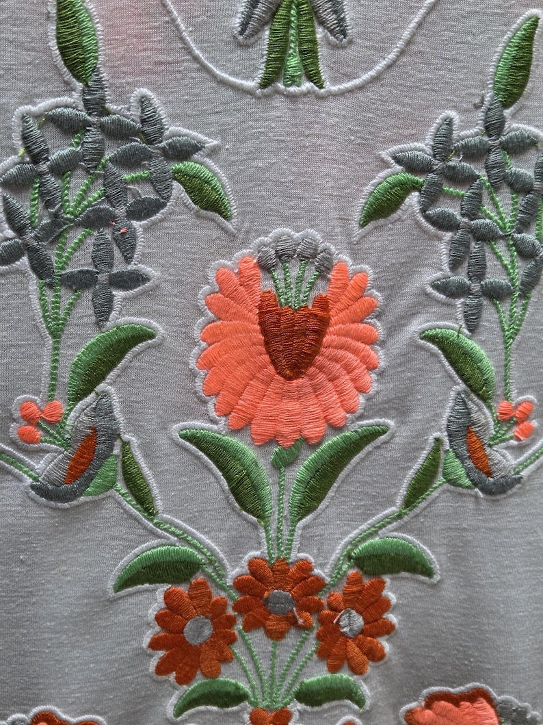 May include: Close-up of a white fabric with intricate floral embroidery. The design features orange, gray, and green flowers and leaves. The embroidery is detailed, with a textured appearance, creating a decorative pattern.