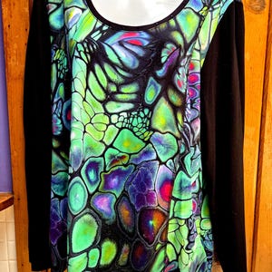 May include: A long-sleeved top with a vibrant, abstract pattern in shades of green, blue, purple, and pink. The sleeves and neckline are solid black. The design features a unique, organic motif, creating a striking visual contrast.