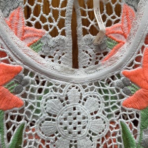 May include: Close-up of a white top with intricate crochet and floral embroidery. The design features orange, green, and gray accents, with a large flower-shaped crochet detail at the center. The top has a keyhole back.