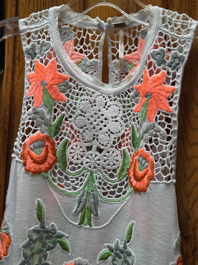 May include: A sleeveless white top with intricate crochet detailing and floral embroidery. The embroidery features orange flowers, green leaves, and gray accents. The top is hanging on a clear hanger.