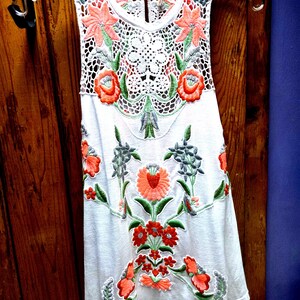 May include: White sleeveless top with floral embroidery in shades of orange, green, and gray. The top features a crochet neckline and floral design. The garment is hanging on a clear hanger against a wooden background.