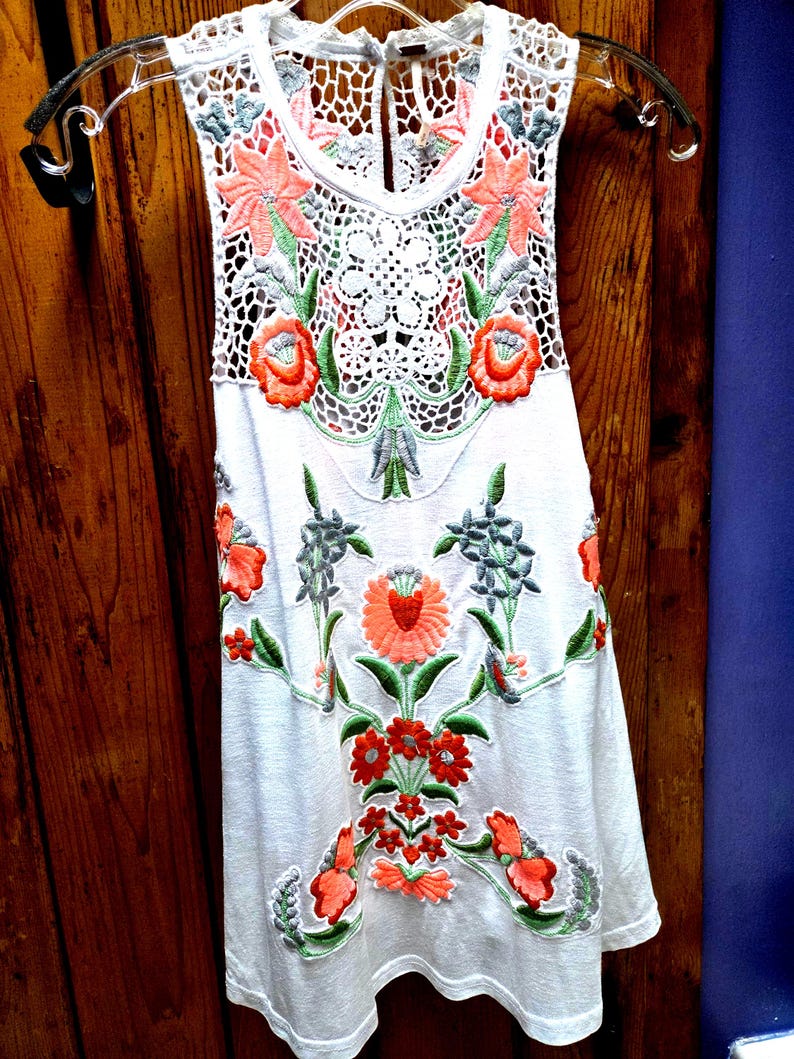 May include: White sleeveless top with floral embroidery in shades of orange, green, and gray. The top features a crochet neckline and floral design. The garment is hanging on a clear hanger against a wooden background.