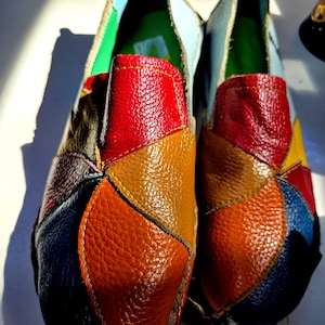 May include: A pair of slip-on shoes constructed from multi-colored, textured leather patches in shades of red, brown, orange, blue, and black. The shoes feature visible stitching along the seams, a light blue interior lining, and a bright green insole.