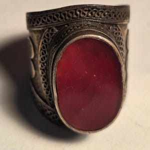 May include: A wide silver ring featuring an oval-shaped, deep red stone set in the center. The metal band is decorated with intricate, fine wire filigree patterns that wrap around the entire circumference of the ring, creating a textured appearance.