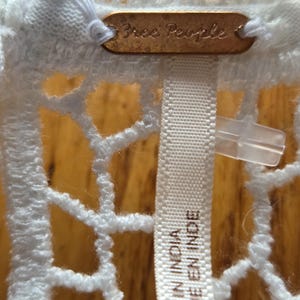 May include: Close-up of a white, open-weave fabric with a tag that reads "Free People." Below the tag is a white fabric label that reads "MADE IN INDIA" and "FABRIQUE EN INDE."