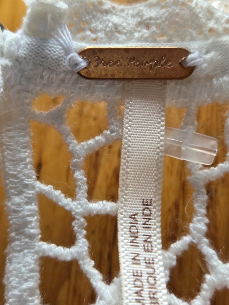 May include: Close-up of a white, open-weave fabric with a tag that reads "Free People." Below the tag is a white fabric label that reads "MADE IN INDIA" and "FABRIQUE EN INDE."