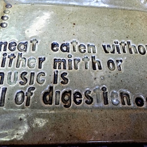 May include: A rectangular ceramic plate with a glossy, speckled light green glaze. The surface features dark, recessed lettering that reads: meat eaten without either mirth or music is ill of digestion, accompanied by small circular indentations.