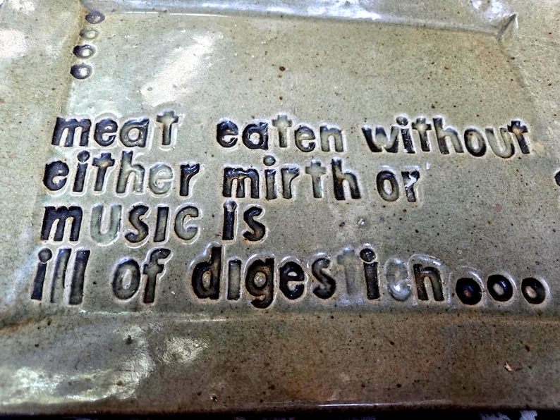 May include: A rectangular ceramic plate with a glossy, speckled light green glaze. The surface features dark, recessed lettering that reads: meat eaten without either mirth or music is ill of digestion, accompanied by small circular indentations.
