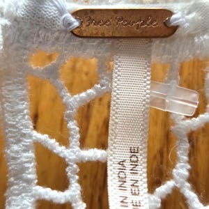 May include: Close-up of a white, open-weave garment with a tag that reads "Free People" in a copper-colored metal. A white fabric tag with the text "MADE IN INDIA / FABRIQUE EN INDE" is also visible.