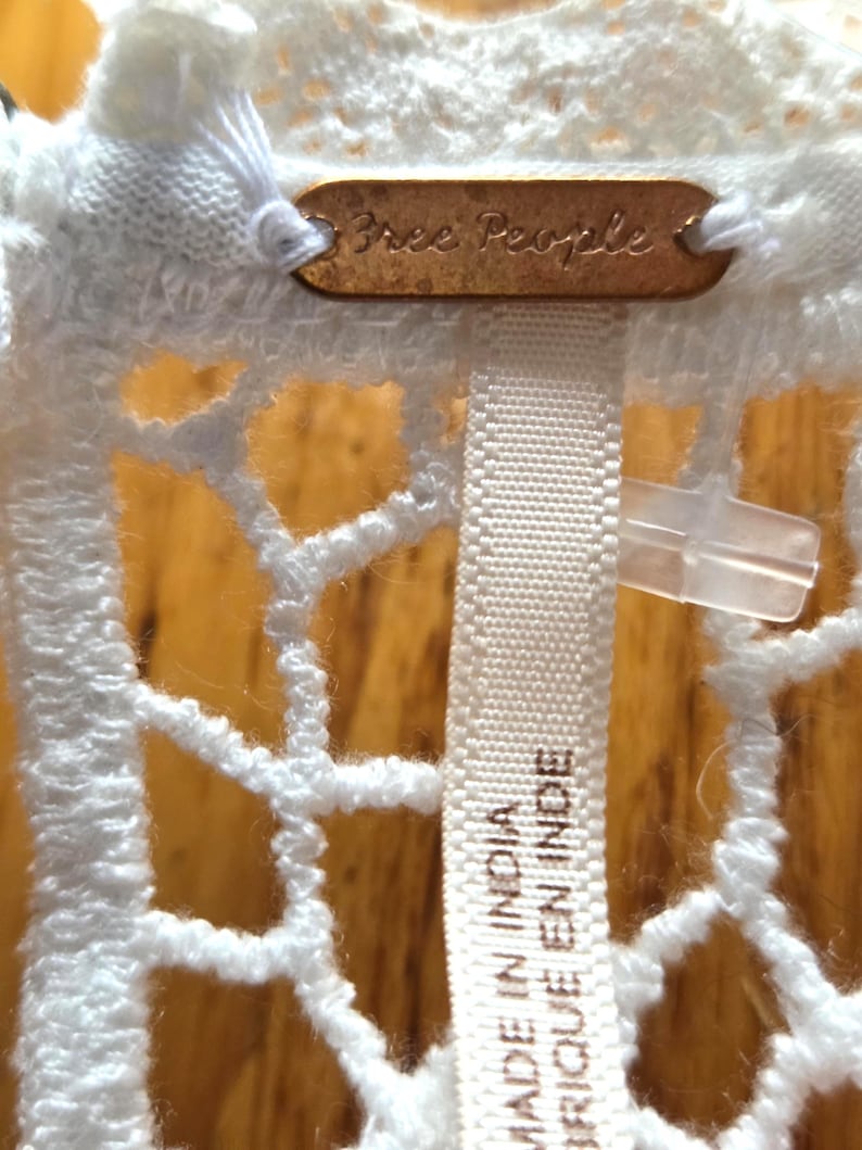 May include: Close-up of a white, open-weave garment with a tag that reads "Free People" in a copper-colored metal. A white fabric tag with the text "MADE IN INDIA / FABRIQUE EN INDE" is also visible.