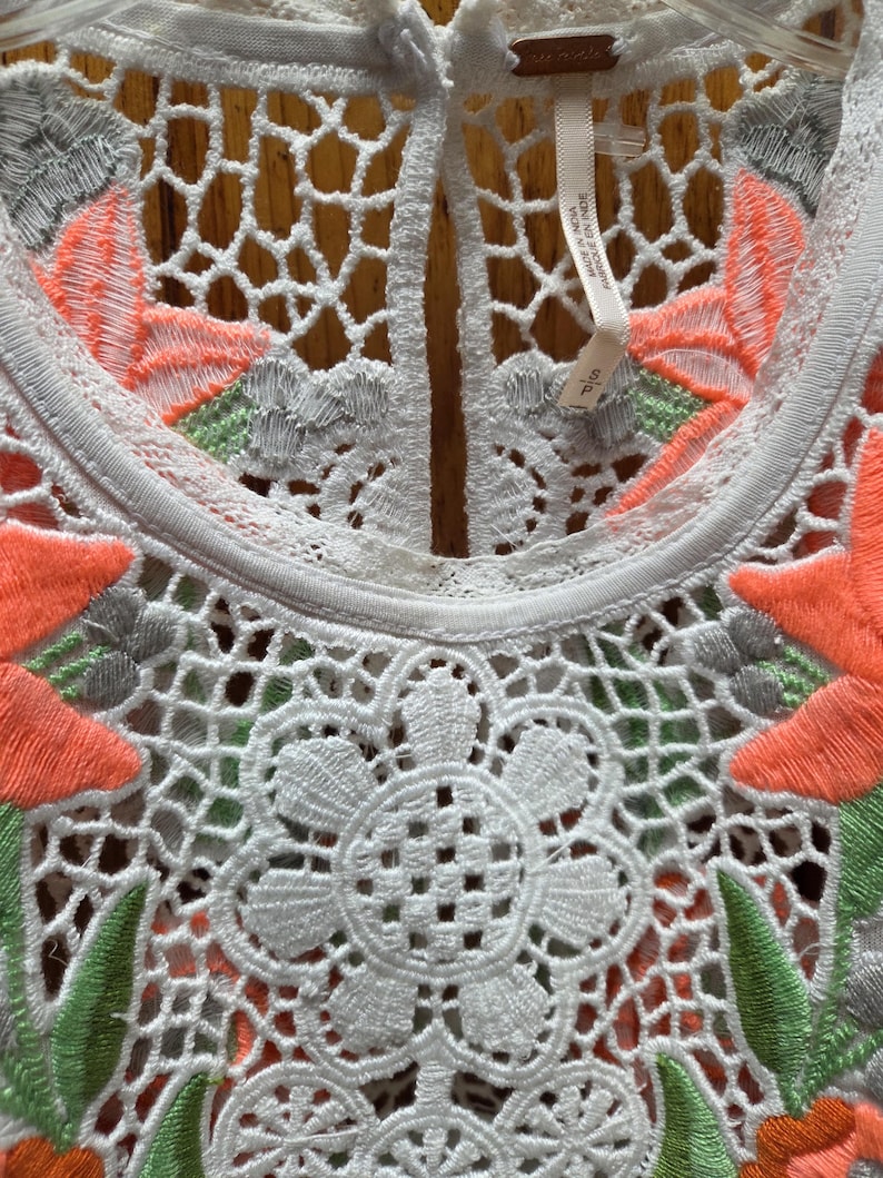 May include: Close-up of a white, embroidered top with a floral design. The top features intricate openwork and is adorned with orange, green, and gray floral embroidery. The tag reads "Free People".